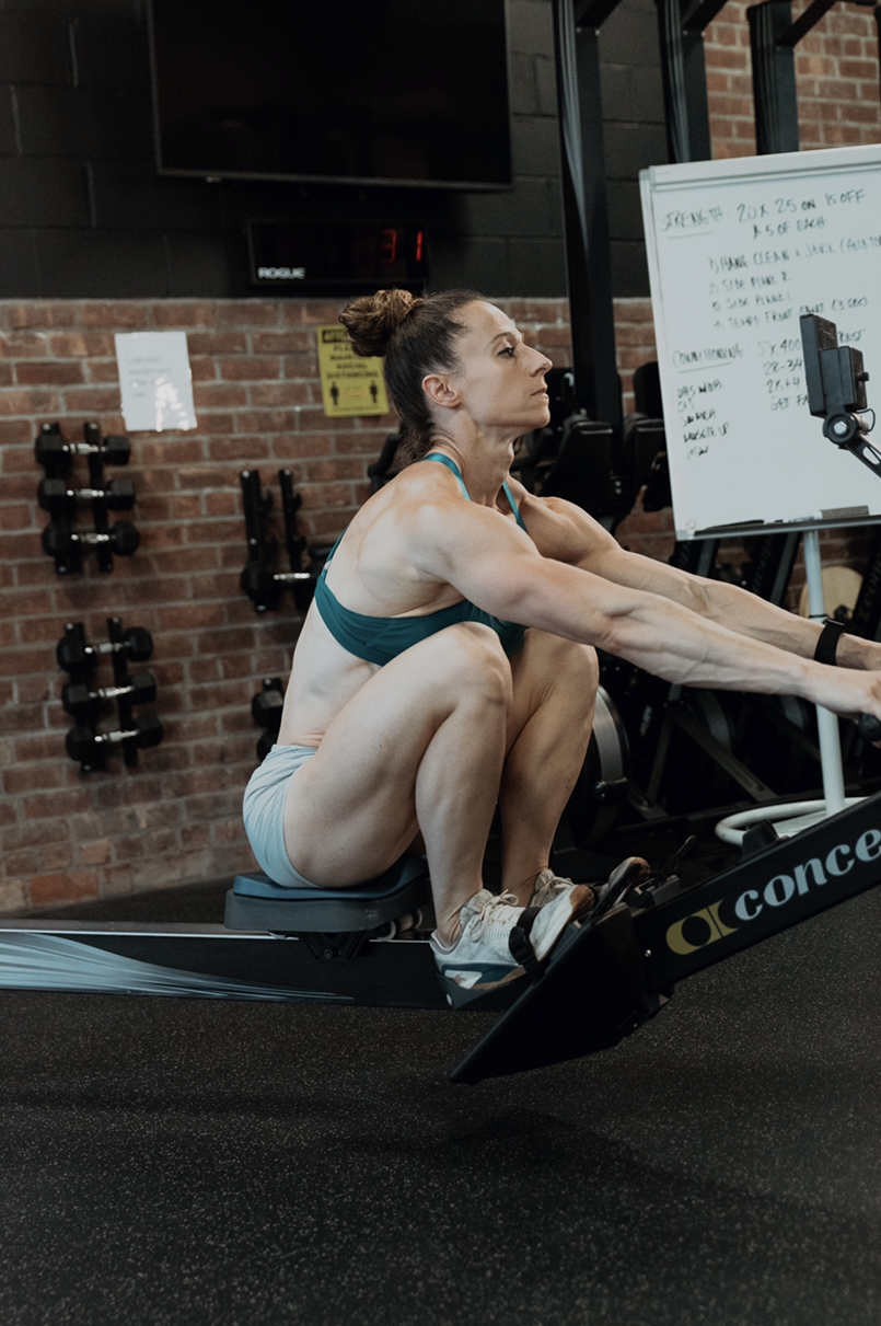 A person exercising on a rowing machine in a gym setting, with workout equipment in the background.