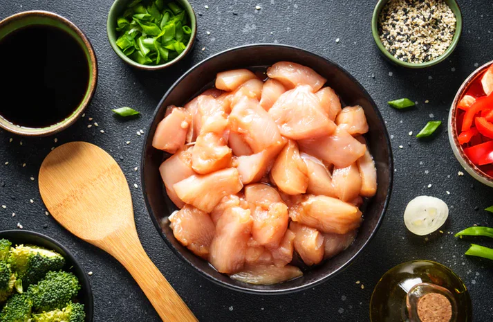 Raw diced chicken breast, chicken starry from New York Prime Beef in a bowl surrounded by fresh vegetables and seasonings, ideal for stir-fry or meal prep.