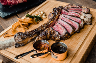 Sliced Tomahawk Ribeye Steak from New York Prime Beef, served on a wooden board with sauces and sides, showcasing premium marbling and flavor.