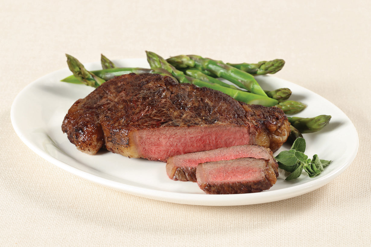 Cooked Boneless Ribeye Steak from New York Prime Beef served with asparagus on a white plate, USDA Prime dry-aged cut with rich flavor.