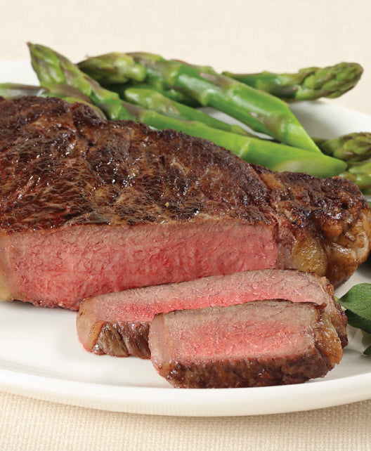 A cooked, juicy boneless ribeye steak with visible marbling, from New York prime beef served on a plate with asparagus in the background.