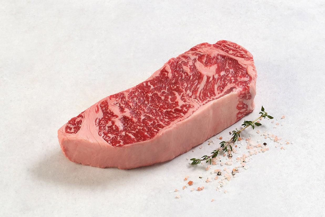 Raw dry aged USDA Prime Boneless New York Strip steak from New York Prime Beef, featuring fine marbling and thickness, displayed on a white background with seasoning