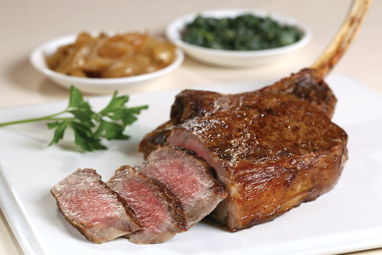 A cooked New York Prime Beef USDA Prime Dry Aged Cowboy Ribeye steak with a golden-brown crust on a plate, accompanied by sliced steak, green vegetables, and a condiment on the side, presented on a white plate with a sprig of parsley.