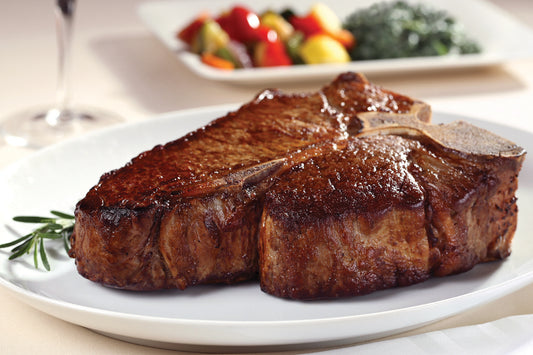 Dry aged USDA Prime Porterhouse steak from New York Prime Beef, cooked to perfection and served on a white plate with vegetables in the background