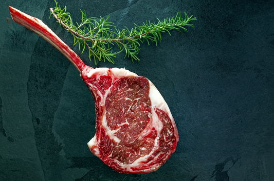 Raw Tomahawk Ribeye Steak from New York Prime Beef with fresh rosemary garnish on a dark stone background, highlighting premium marbling.