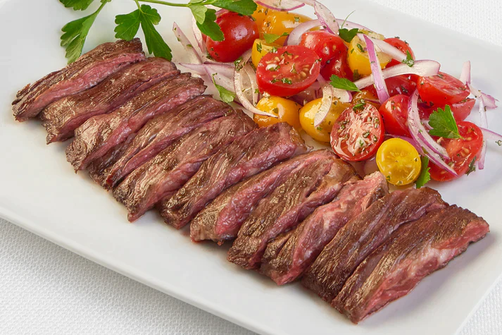 Sliced Skirt Steak from New York Prime Beef served on a white plate with fresh tomato and onion salad, tender and flavorful cut.