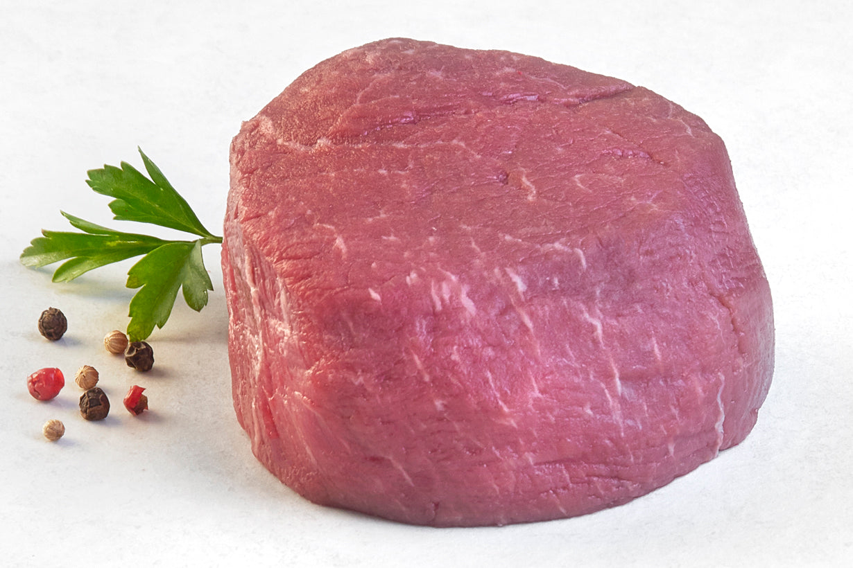 A single, uncooked grass-fed filet Mignon steak on a white plate with a sprig of parsley to the side.
