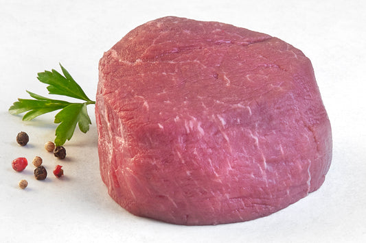 A single, uncooked grass-fed filet Mignon steak on a white plate with a sprig of parsley to the side.